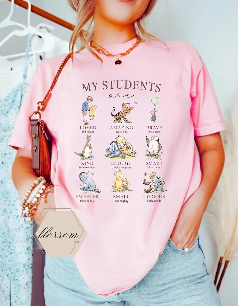 May include: Pink t-shirt with the text "MY STUDENTS are" and illustrations of Winnie the Pooh characters with positive adjectives like "LOVED" and "AMAZING". The shirt also has the word "blossom" printed on it.