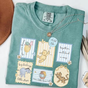 May include: A light green Comfort Colors t-shirt featuring cartoon illustrations of Winnie the Pooh characters. The shirt has several framed images with text, including "you are brave," "child life," and "together we'll find a way."