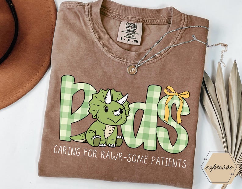 May include: A brown Comfort Colors t-shirt featuring the word "Peds" in a green and white plaid design, with a cartoon triceratops. The text "CARING FOR RAWR-SOME PATIENTS" is below. A silver necklace is on the shirt. The shirt is a size S-P-CH.