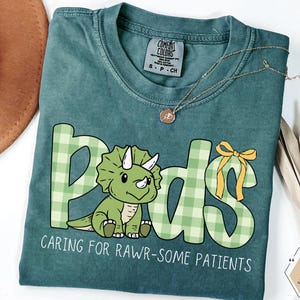 May include: A teal Comfort Colors t-shirt with the word "Pods" in a green plaid design and a cartoon triceratops. The text "Caring for Rawr-some Patients" is below the graphic. A gold necklace is draped across the shirt.