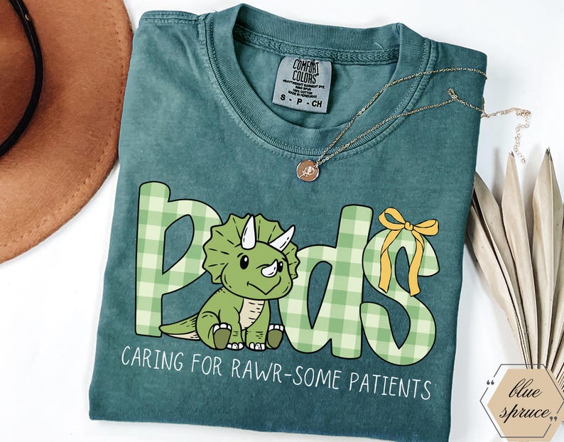 May include: A teal Comfort Colors t-shirt with the word "Pods" in a green plaid design and a cartoon triceratops. The text "Caring for Rawr-some Patients" is below the graphic. A gold necklace is draped across the shirt.