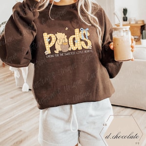 May include: A brown sweatshirt with the word "Pods" in yellow gingham print, featuring a cartoon bear and the text "Caring for the Sweetest Little Hearts." The sweatshirt is paired with gray shorts.
