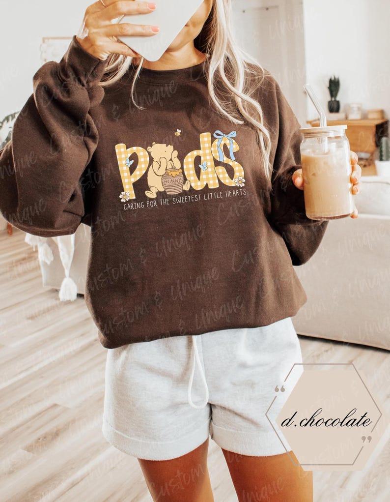 May include: A brown sweatshirt with the word "Pods" in yellow gingham print, featuring a cartoon bear and the text "Caring for the Sweetest Little Hearts." The sweatshirt is paired with gray shorts.