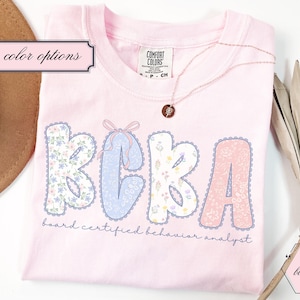 May include: A pale pink t-shirt featuring the letters BCBA in a floral design. The text "board certified behavior analyst" is below the letters. A gold necklace with a pendant is on the shirt.