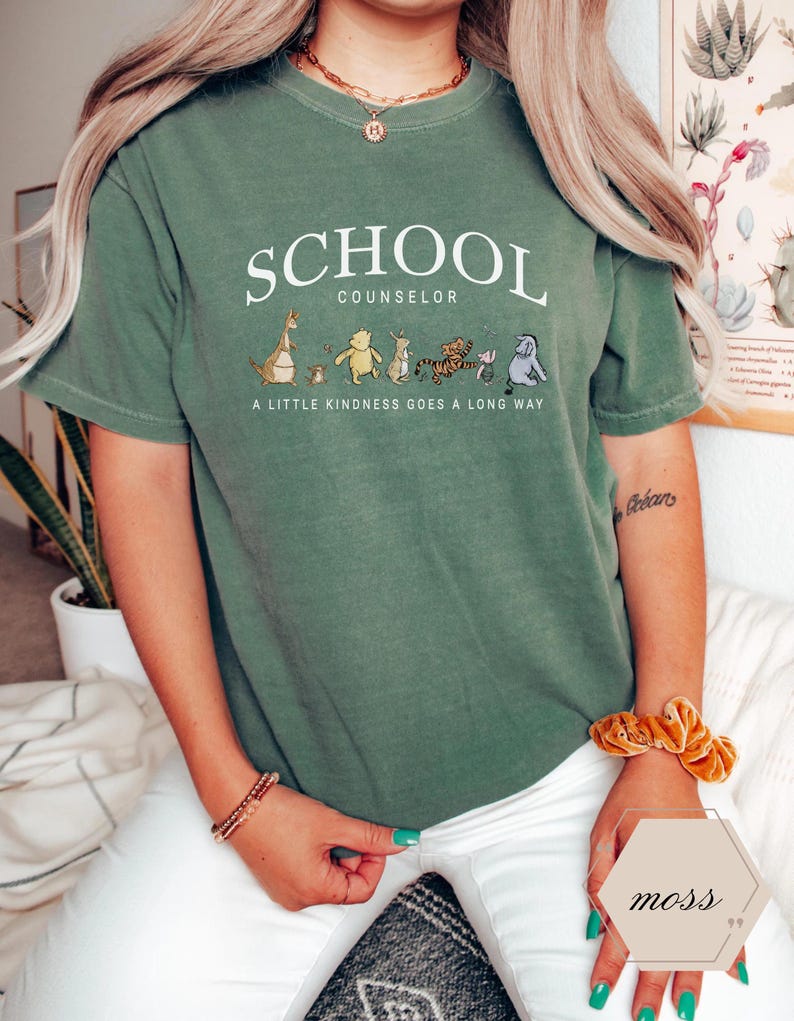 May include: A moss green short-sleeve t-shirt featuring the white text SCHOOL COUNSELOR and the phrase A LITTLE KINDNESS GOES A LONG WAY, accompanied by a graphic illustration of six cartoon characters walking in a line across the chest.