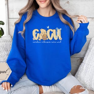 May include: A royal blue sweatshirt with the letters "CCU" in a yellow gingham pattern, featuring a cartoon bear and a butterfly. The text "cardiac intensive care unit" is below the letters. A white and gold logo is in the lower left corner.