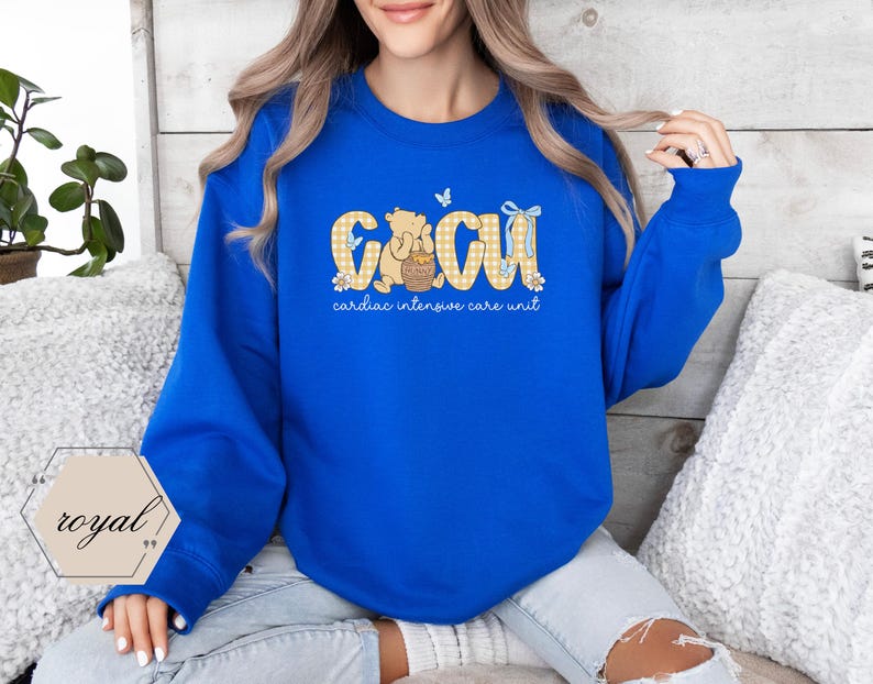 May include: A royal blue sweatshirt with the letters "CCU" in a yellow gingham pattern, featuring a cartoon bear and a butterfly. The text "cardiac intensive care unit" is below the letters. A white and gold logo is in the lower left corner.