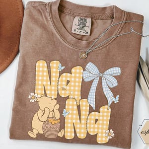 以下が含まれることがあります： 黄色のギンガムチェック柄で「Na Na」の文字が入った茶色のコンフォートカラーズTシャツ。くまのプーさんのグラフィック、青いリボン、蝶がシャツを飾っています。ペンダント付きのシルバーネックレスがシャツの上にあります。