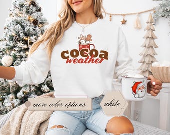 May include: White sweatshirt featuring the text "cocoa weather" in brown and red, with a hot cocoa mug design. The image also shows a white mug with a Santa Claus graphic. The sweatshirt is part of a winter-themed collection.