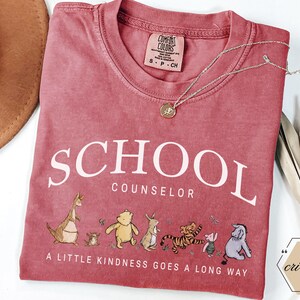 May include: A dusty rose crewneck t-shirt featuring the white text SCHOOL COUNSELOR and the phrase A LITTLE KINDNESS GOES A LONG WAY. Below the text is a graphic of various cartoon animals including a bear, a tiger, a donkey, and a kangaroo.