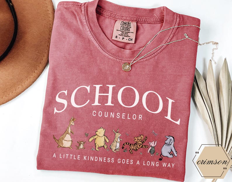 May include: A dusty rose crewneck t-shirt featuring the white text SCHOOL COUNSELOR and the phrase A LITTLE KINDNESS GOES A LONG WAY. Below the text is a graphic of various cartoon animals including a bear, a tiger, a donkey, and a kangaroo.