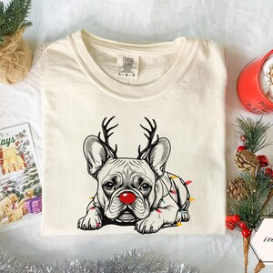 May include: Ivory-colored t-shirt featuring a French bulldog with reindeer antlers and a red nose, adorned with Christmas lights. The shirt has a crew neck and a tag that reads "Comfort Colors". A holiday card and a red mug are in the background.