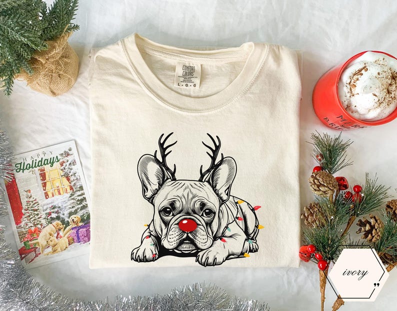 May include: Ivory-colored t-shirt featuring a French bulldog with reindeer antlers and a red nose, adorned with Christmas lights. The shirt has a crew neck and a tag that reads "Comfort Colors". A holiday card and a red mug are in the background.