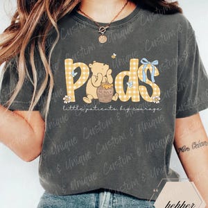 May include: Dark gray t-shirt featuring the word "Pods" in yellow plaid letters, with a cartoon bear illustration. The shirt also includes the text "little patients, big courage". A blue bow and floral accents complete the design.