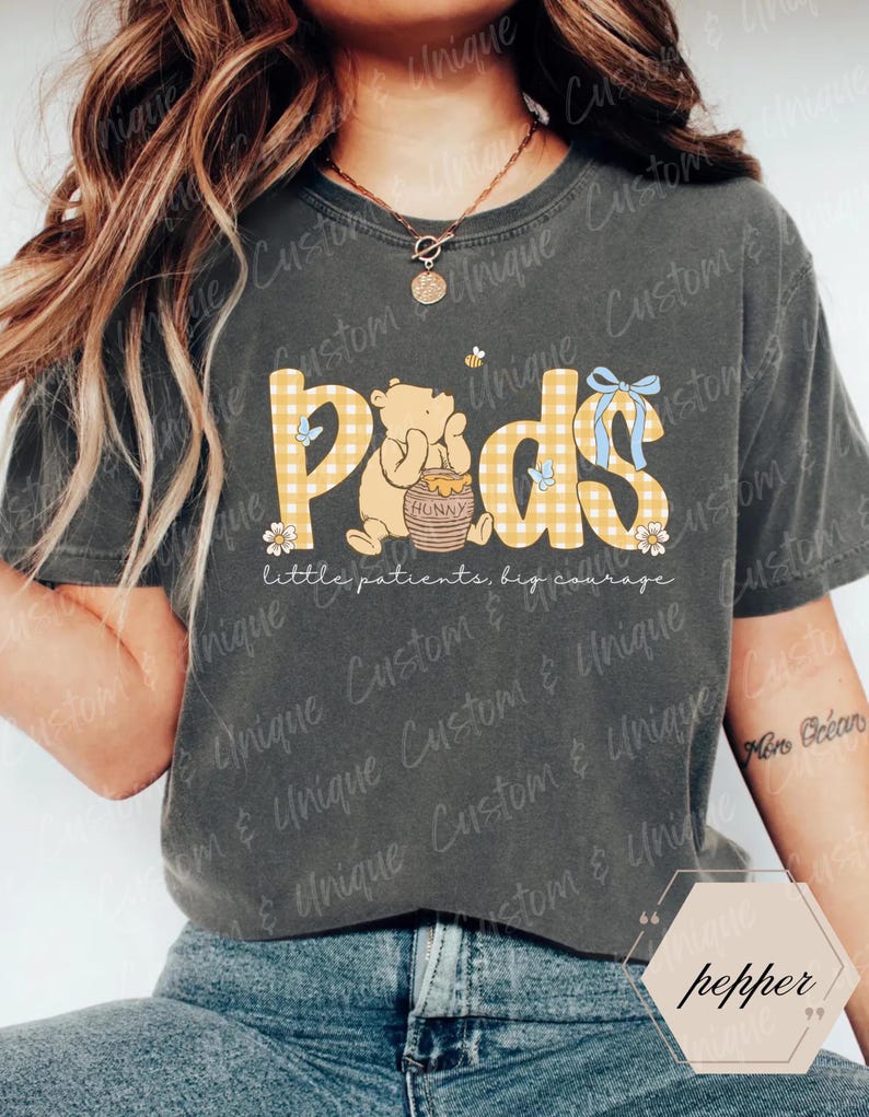 May include: Dark gray t-shirt featuring the word "Pods" in yellow plaid letters, with a cartoon bear illustration. The shirt also includes the text "little patients, big courage". A blue bow and floral accents complete the design.