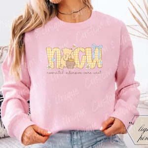 May include: Light pink sweatshirt with the letters "NICU" in yellow plaid, featuring a bear holding a honey pot. The text "neonatal intensive care unit" is below the letters. A blue bow and a bee are also part of the design.