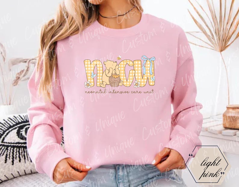 May include: Light pink sweatshirt with the letters "NICU" in yellow plaid, featuring a bear holding a honey pot. The text "neonatal intensive care unit" is below the letters. A blue bow and a bee are also part of the design.