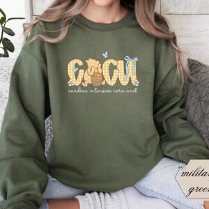 May include: Military green sweatshirt featuring the text "CICU cardiac intensive care unit" in yellow gingham lettering. The design includes a cartoon bear, butterflies, and floral accents. A casual, comfortable style.