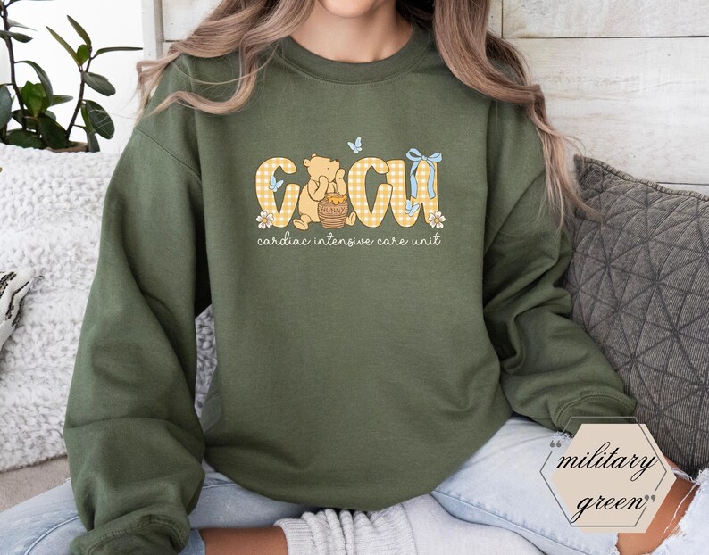 May include: Military green sweatshirt featuring the text "CICU cardiac intensive care unit" in yellow gingham lettering. The design includes a cartoon bear, butterflies, and floral accents. A casual, comfortable style.