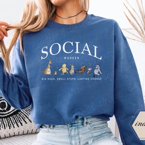 Pu&ograve; includere: Una felpa blu con la scritta bianca "SOCIAL WORKER" a forma di arco. Sotto, personaggi dei cartoni animati. La frase "BIG HUGS, SMALL STEPS, LASTING CHANGE" &egrave; stampata sotto.