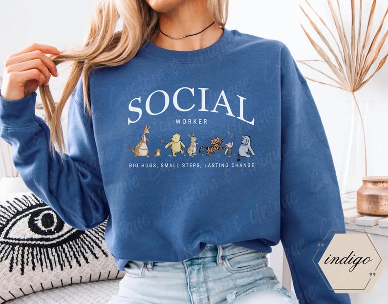 Pu&ograve; includere: Una felpa blu con la scritta bianca "SOCIAL WORKER" a forma di arco. Sotto, personaggi dei cartoni animati. La frase "BIG HUGS, SMALL STEPS, LASTING CHANGE" &egrave; stampata sotto.