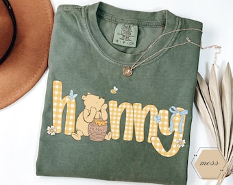 May include: Green t-shirt with the word "hunny" in yellow gingham letters, featuring a cartoon bear, honey pot, bees, and butterflies. A gold necklace with a pendant rests on the shirt. The shirt is a Comfort Colors brand, size S.