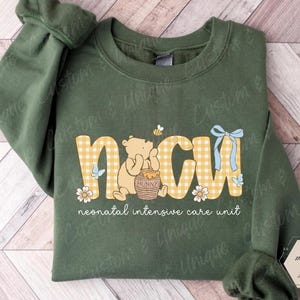 May include: Military green sweatshirt with the letters "NICU" in a yellow gingham pattern, featuring a cartoon bear, honey pot, and flowers. The text "neonatal intensive care unit" is below the letters.