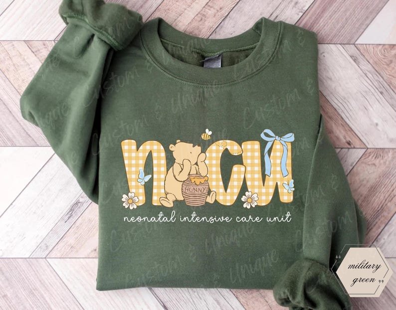 May include: Military green sweatshirt with the letters "NICU" in a yellow gingham pattern, featuring a cartoon bear, honey pot, and flowers. The text "neonatal intensive care unit" is below the letters.