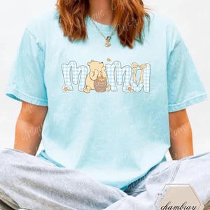 May include: Light blue t-shirt with the word "Mama" in a blue gingham pattern. A cartoon bear holding a honey pot is next to the word "Mama". A yellow bow and small flowers are also part of the design.