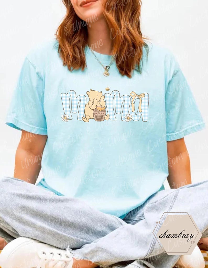 May include: Light blue t-shirt with the word "Mama" in a blue gingham pattern. A cartoon bear holding a honey pot is next to the word "Mama". A yellow bow and small flowers are also part of the design.