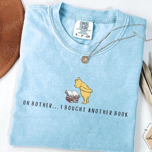 May include: Light blue Comfort Colors t-shirt with the text "OH BOTHER... I BOUGHT ANOTHER BOOK" and a cartoon bear illustration. A silver necklace and a brown hat are also visible.