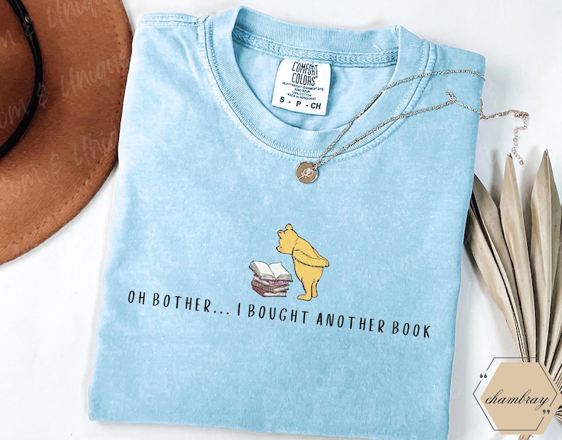 May include: Light blue Comfort Colors t-shirt with the text "OH BOTHER... I BOUGHT ANOTHER BOOK" and a cartoon bear illustration. A silver necklace and a brown hat are also visible.