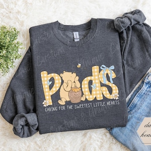 May include: Dark heather gray sweatshirt with the word "Pads" in yellow gingham letters, featuring a cartoon bear and a honey pot. The text "Caring for the sweetest little hearts" is below the design.