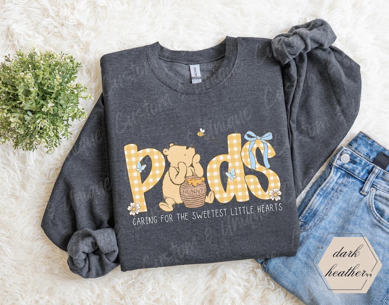 May include: Dark heather gray sweatshirt with the word "Pads" in yellow gingham letters, featuring a cartoon bear and a honey pot. The text "Caring for the sweetest little hearts" is below the design.