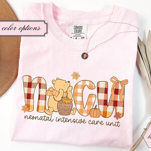 May include: Light pink t-shirt with the letters "NICU" in a plaid pattern, featuring a cartoon bear, pumpkin, and leaves. The text "neonatal intensive care unit" is below the letters. A necklace and a hat are also visible.
