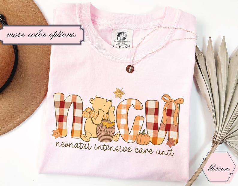 May include: Light pink t-shirt with the letters "NICU" in a plaid pattern, featuring a cartoon bear, pumpkin, and leaves. The text "neonatal intensive care unit" is below the letters. A necklace and a hat are also visible.