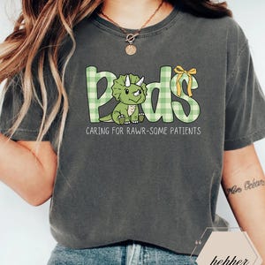 May include: Dark grey t-shirt with the word "Peds" in green plaid letters, featuring a cartoon triceratops. The text "Caring for Rawr-some Patients" is below the word "Peds". A yellow bow is on the letter "s".
