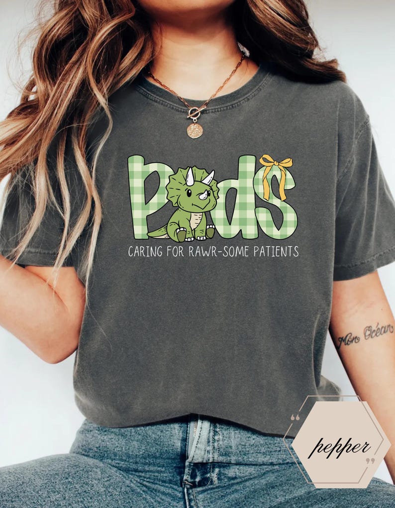 May include: Dark grey t-shirt with the word "Peds" in green plaid letters, featuring a cartoon triceratops. The text "Caring for Rawr-some Patients" is below the word "Peds". A yellow bow is on the letter "s".