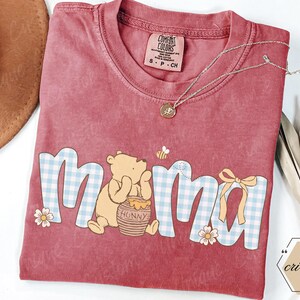 May include: A red Comfort Colors t-shirt with the word "mama" in a blue and white gingham pattern, featuring Winnie the Pooh and a honey pot. A gold necklace and a small bee graphic are also visible.
