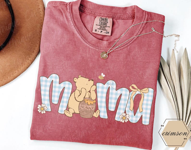 May include: A red Comfort Colors t-shirt with the word "mama" in a blue and white gingham pattern, featuring Winnie the Pooh and a honey pot. A gold necklace and a small bee graphic are also visible.