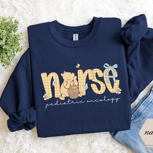 Peut inclure: Sweat-shirt bleu marine avec le mot "nurse" en imprim&eacute; vichy jaune, repr&eacute;sentant un ours de dessin anim&eacute; tenant un pot de miel. Le texte "pediatric oncology" est en dessous.