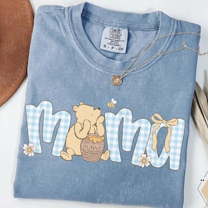 Op de afbeelding: Blauw Comfort Colors T-shirt met het woord "mama" in een lichtblauw ruitpatroon. Een Winnie de Poeh beer houdt een honingpot vast. Een gouden ketting ligt op het shirt.