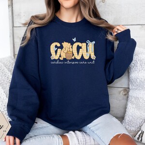 May include: Navy blue sweatshirt with the letters "CICU" in a yellow gingham pattern, featuring a cartoon bear holding a honey pot. The words "cardiac intensive care unit" are printed below the letters. A butterfly and bow accent the design.