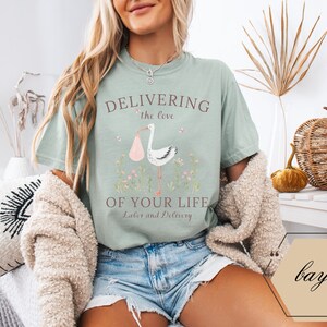 Op de afbeelding: Een lichtblauw t-shirt met een afbeelding van een ooievaar die een baby draagt. De tekst op het shirt luidt "Delivering the love of your life Labor and Delivery".