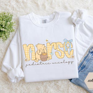 Peut inclure: Sweat-shirt blanc avec le mot "nurse" en lettres &agrave; carreaux jaunes, avec un motif Winnie l'ourson. Le motif comprend un pot de miel, des abeilles et le texte "pediatric oncology". Un n&oelig;ud bleu clair orne le motif.