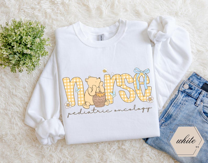 Peut inclure: Sweat-shirt blanc avec le mot "nurse" en lettres &agrave; carreaux jaunes, avec un motif Winnie l'ourson. Le motif comprend un pot de miel, des abeilles et le texte "pediatric oncology". Un n&oelig;ud bleu clair orne le motif.