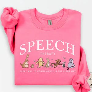 May include: A bright pink sweatshirt with the words "SPEECH THERAPY" in white arched text. Below the text are cartoon illustrations of Winnie the Pooh characters. The phrase "EVERY WAY TO COMMUNICATE IS THE RIGHT WAY" is printed below the illustrations.