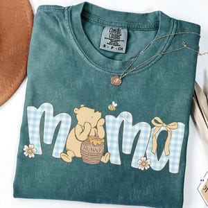 May include: Teal Comfort Colors t-shirt with the word "mama" in a blue gingham pattern, featuring a Winnie the Pooh graphic. A gold necklace and a brown hat are also visible.