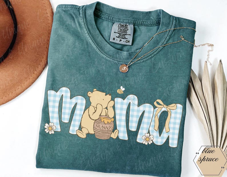 May include: Teal Comfort Colors t-shirt with the word "mama" in a blue gingham pattern, featuring a Winnie the Pooh graphic. A gold necklace and a brown hat are also visible.