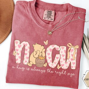 May include: A maroon Comfort Colors t-shirt featuring the word "N CU" in a playful font with heart patterns. A cartoon bear sits beside a honey pot. The text "a hug is always the right size" is below. A gold necklace and a hat are also visible.